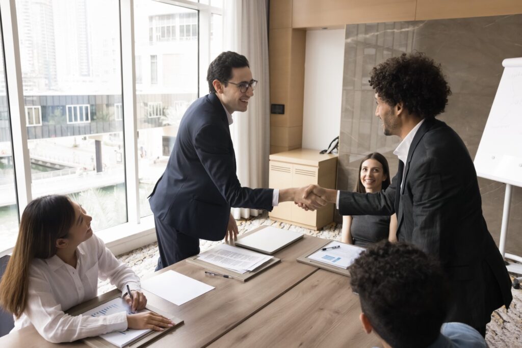 Man in suit shaking another mans hand over a table with 3 other people sitting at the table