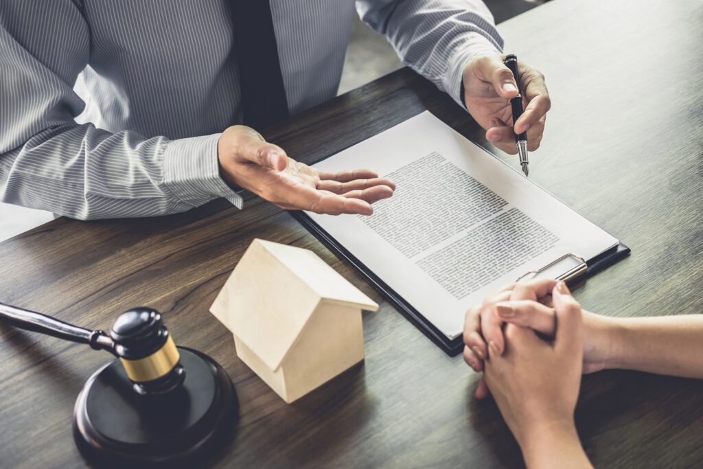 An attorney sitting across from a client at a desk in front of paperwork and next to a gavel and small model house.